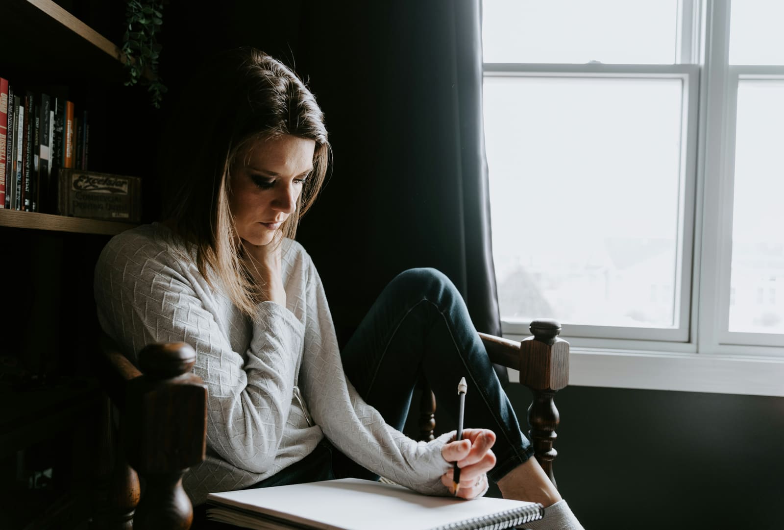 A woman sitting alone by a window in a quiet, softly lit room, writing in a notebook.