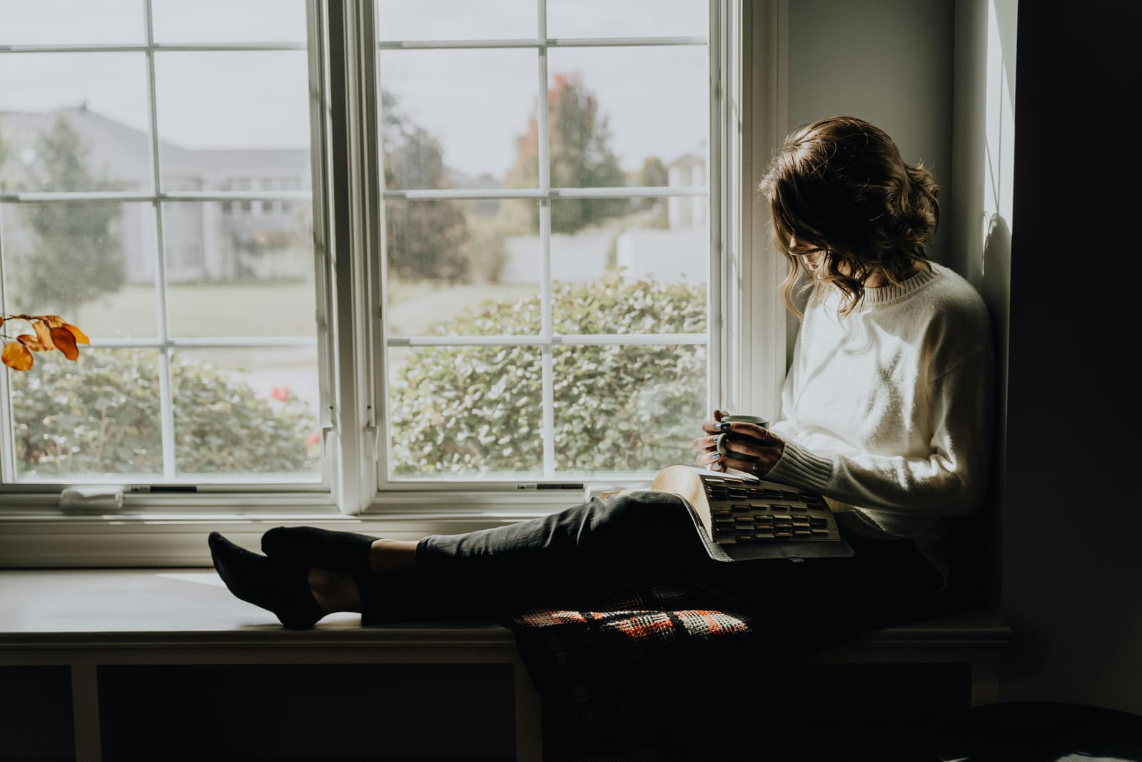 A woman sitting on a window bench with her feet up, an open tabbed Bible in her lap and a mug of coffee in hand, cloudy daylight outside.