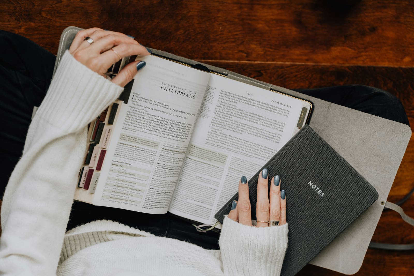 An overhead view of hands resting on an open Bible beside a notebook labelled Notes, on a wooden desk.