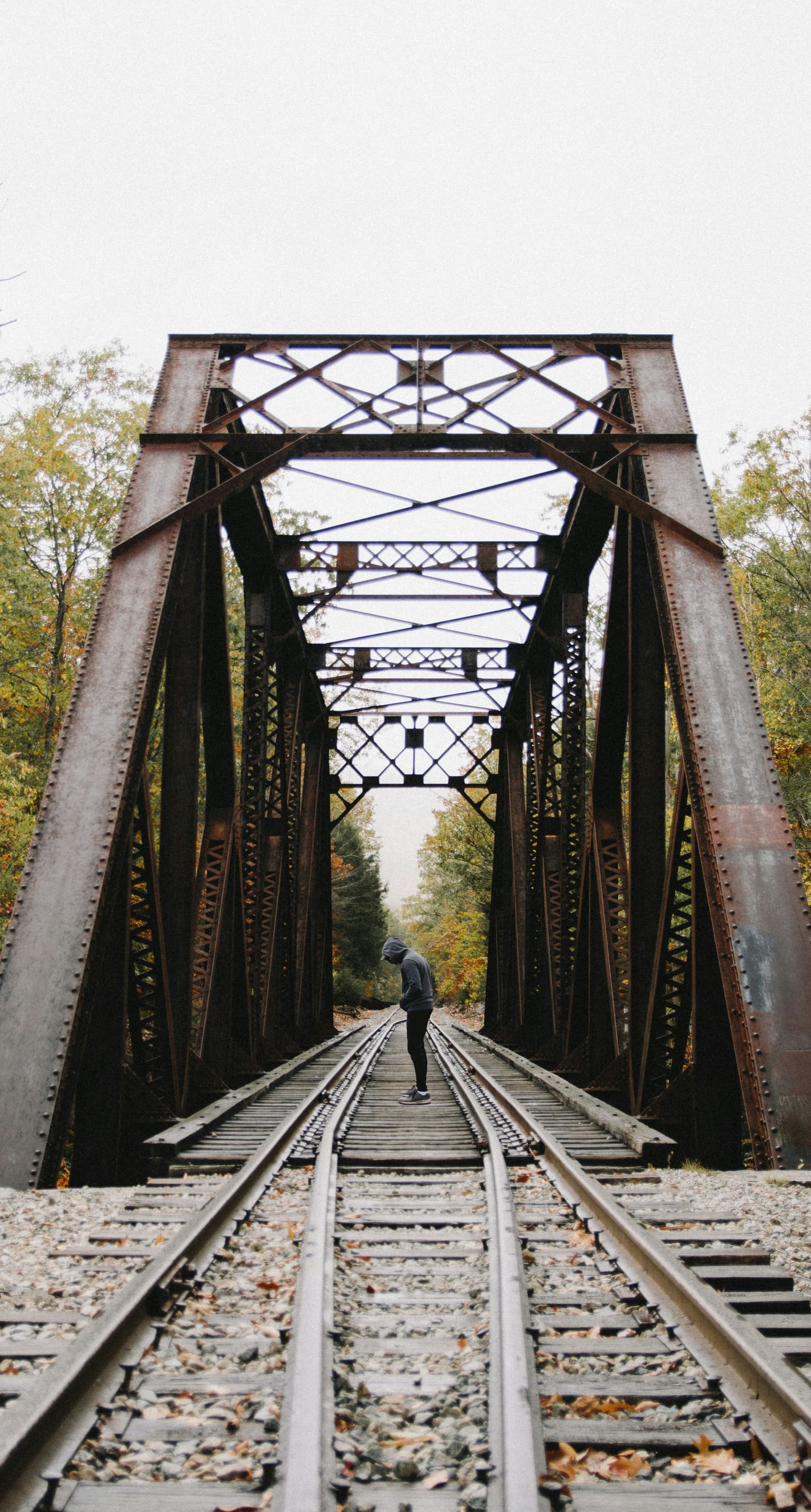 A lone figure standing beneath an old iron railway bridge, autumn trees on either side, head bowed.