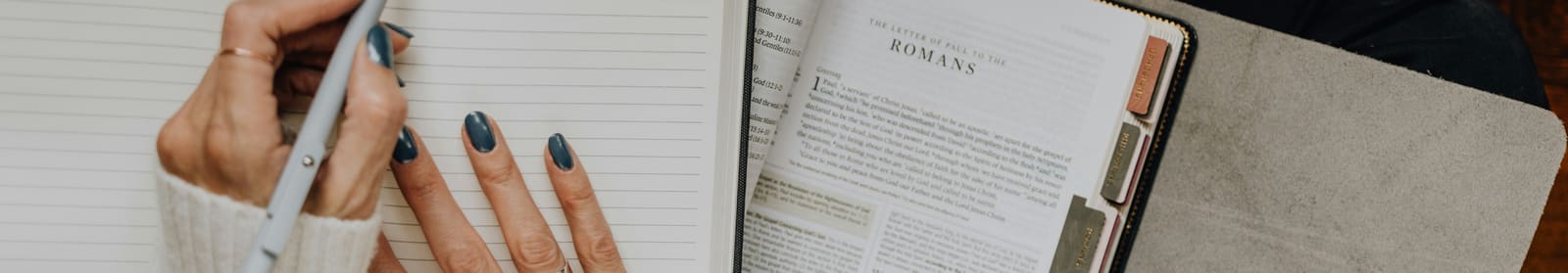 Overhead view of a hand writing in a journal beside an open, tabbed Bible turned to Romans.