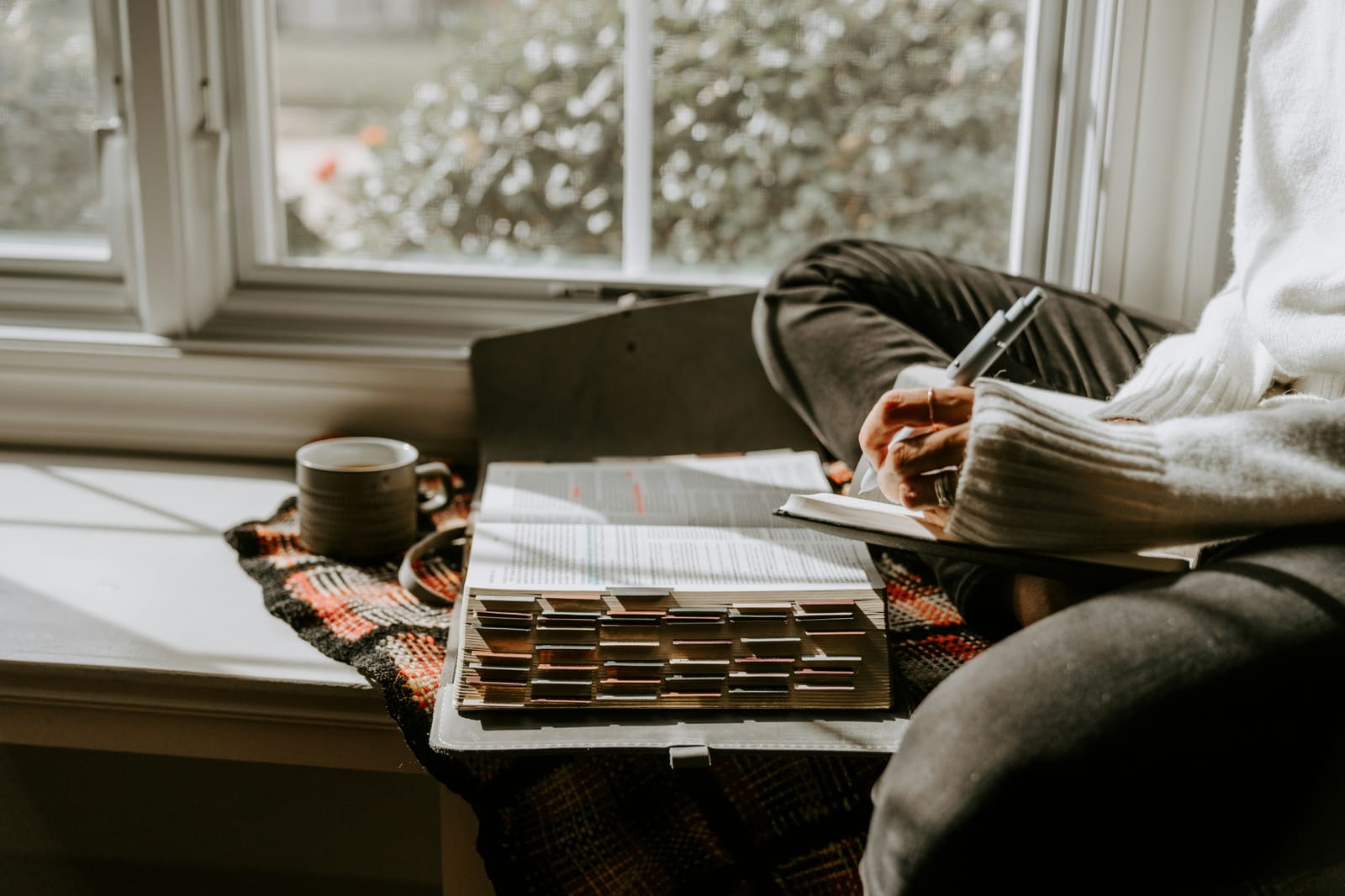 A person marking a passage in an open Bible beside a window, with a cup of coffee close by, in daylight.