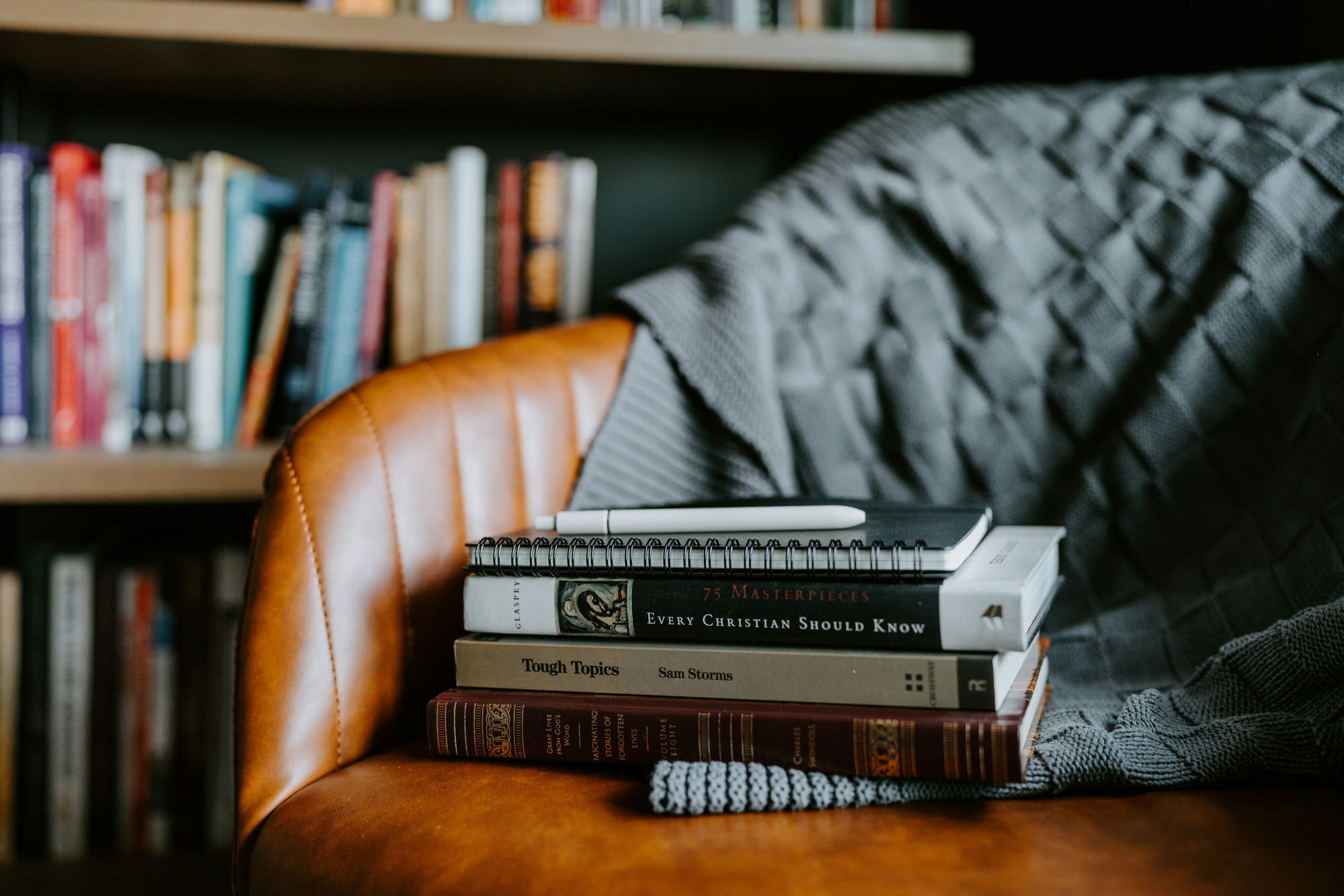 A stack of well-loved Christian books and a notebook resting on a leather armchair, with a bookshelf in the background.
